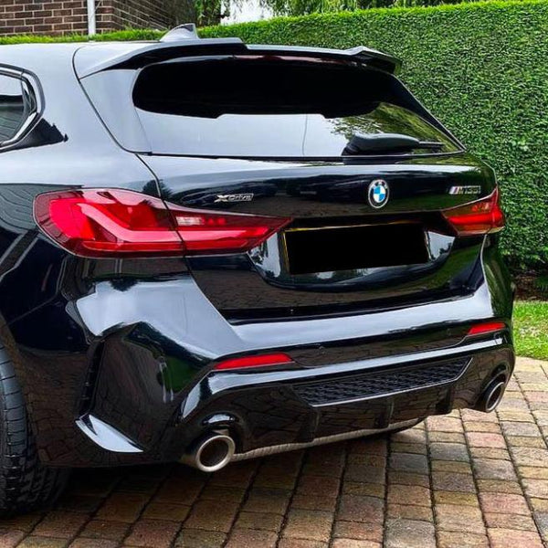 A shiny black BMW X5M parked on a brick driveway, surrounded by a brick wall and trimmed hedge, with visible red taillights and dual exhaust pipes.