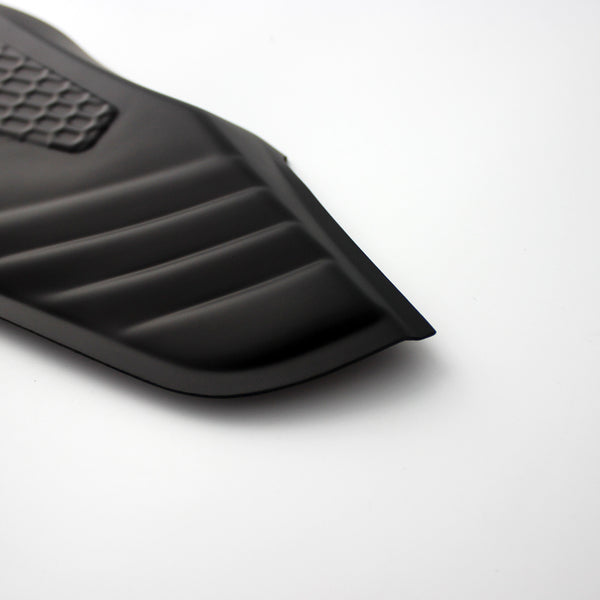 A black, ribbed car floor mat corner with a textured rectangular section rests on a smooth white surface, showing its contoured edges.