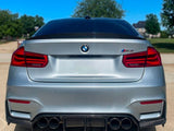 A silver BMW M3 with quad exhaust tips is parked on a driveway. The background features suburban homes and trees under a clear blue sky.