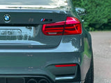 A gray BMW M3 is parked on a street. The rear features red taillights and dual exhausts, surrounded by greenery and a paved road.