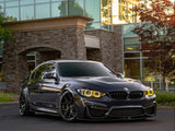 A sleek, dark gray BMW sedan is parked on smooth pavement. The car's headlights glow yellow, and it's situated in front of a modern building with large windows and stone walls.