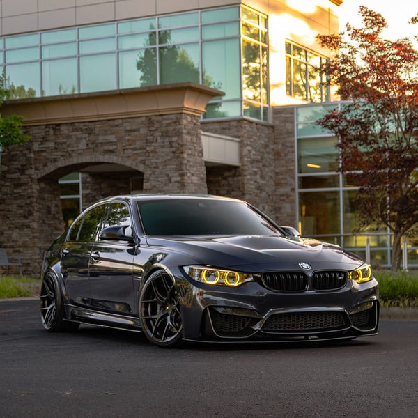 A sleek, dark gray BMW sedan is parked on smooth pavement. The car's headlights glow yellow, and it's situated in front of a modern building with large windows and stone walls.