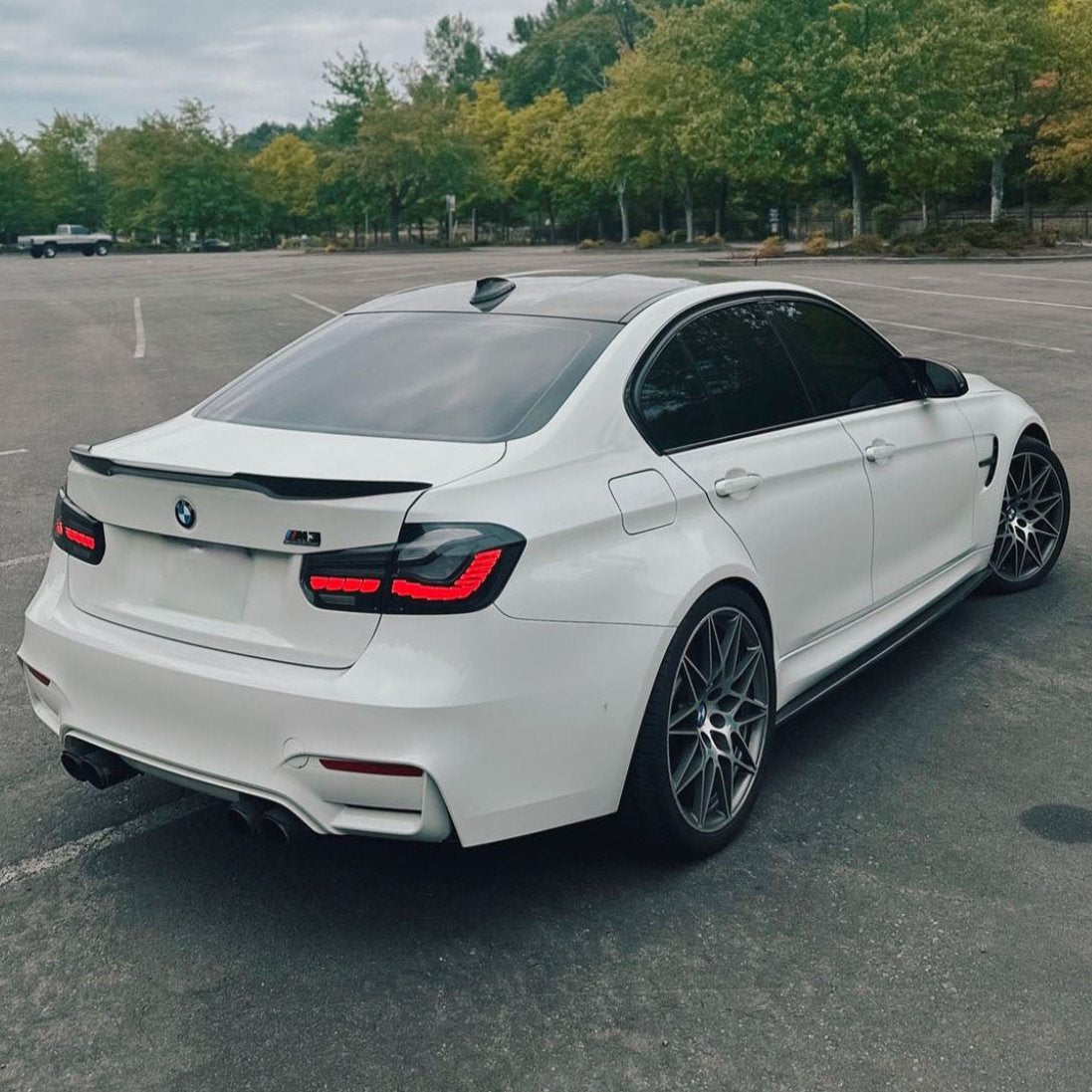 A white BMW M3 is parked in an empty parking lot, surrounded by green trees under a cloudy sky.