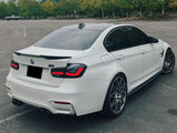A white BMW M3 is parked in an empty lot. The car features tinted windows, dual exhausts, and distinctive alloy wheels. Surrounding the lot are lush green trees and overcast skies.