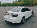 A white BMW sports sedan is parked in an empty lot. It features tinted windows, a rear spoiler, dual exhausts, and dark wheels. Green trees line the background under a cloudy sky.