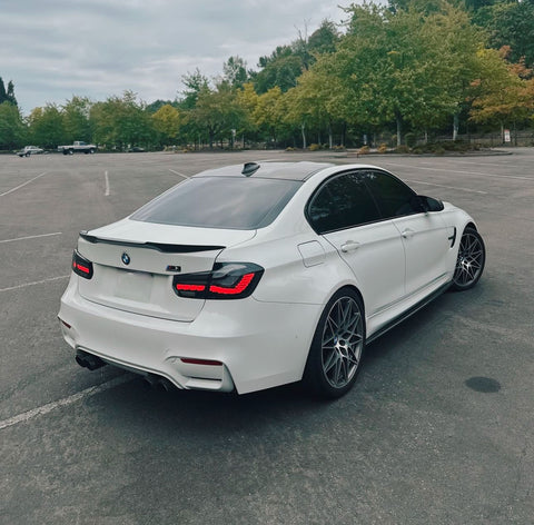 A white BMW sports sedan is parked in an empty lot. It features tinted windows, a rear spoiler, dual exhausts, and dark wheels. Green trees line the background under a cloudy sky.