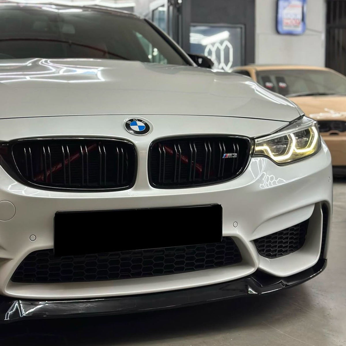 A white BMW car with illuminated headlights is parked indoors; the front grille features the BMW logo and "M3" badge. Another car is partially visible in the background.