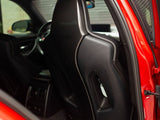A black, sleek car seat with carbon fiber detailing is positioned inside a red vehicle, visible through the open rear door, with the steering wheel and dashboard in the background.