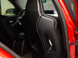 A sleek, black leather car seat with a carbon fiber backrest is positioned inside a red vehicle, surrounded by a modern interior featuring rear climate control and sleek design elements.
