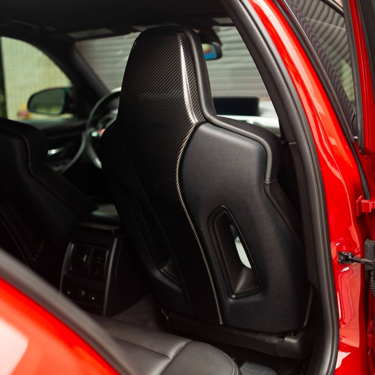 A sleek, black leather car seat with a carbon fiber backrest is positioned inside a red vehicle, surrounded by a modern interior featuring rear climate control and sleek design elements.
