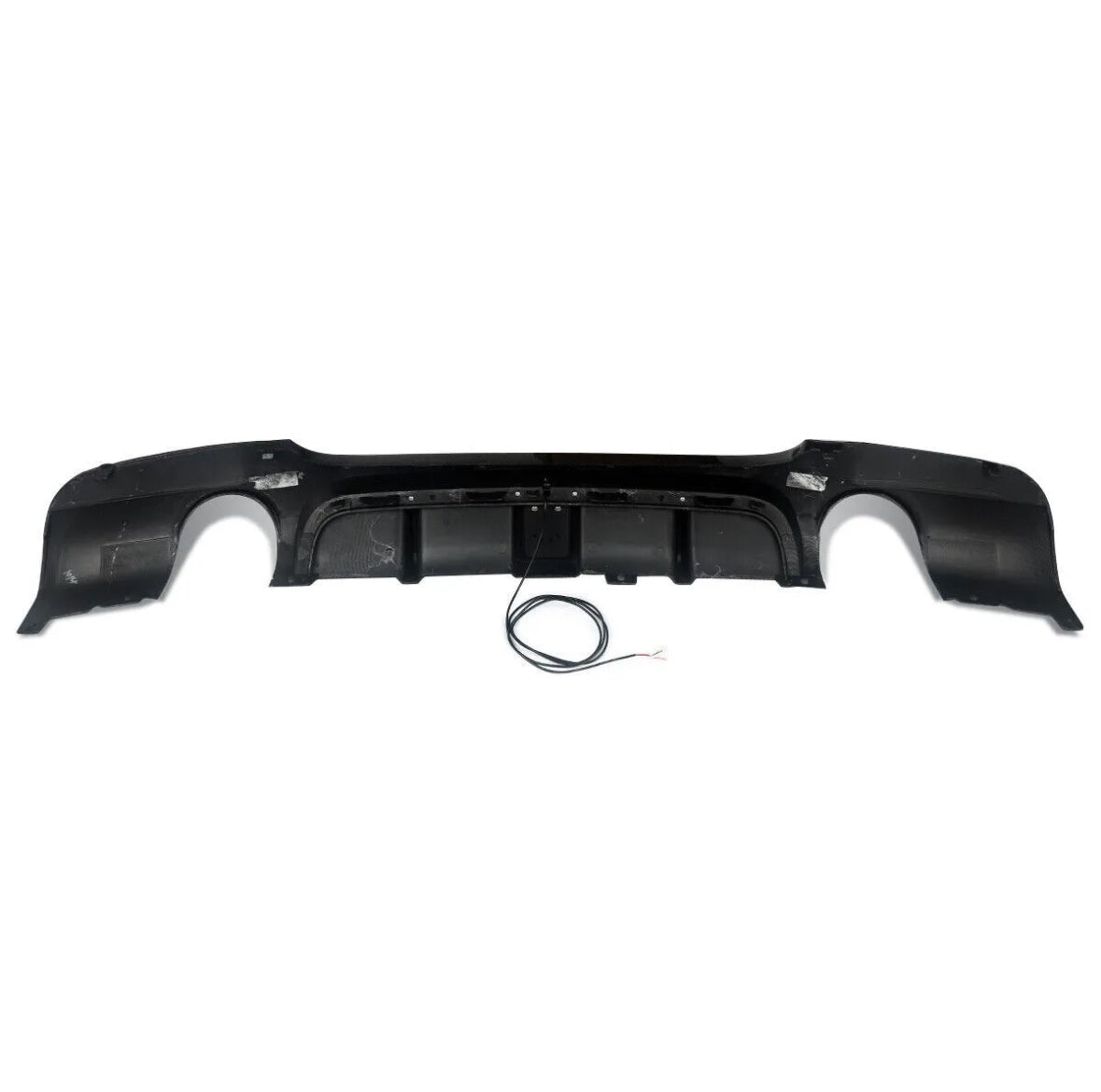 A black car bumper diffuser with integrated exhaust cutouts sits on a white background. A coiled black wire with exposed ends lies beneath it.