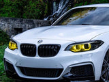 A white BMW with illuminated yellow headlights is parked on grass. Behind it, a stone wall and a small waterfall surrounded by greenery provide a scenic background.