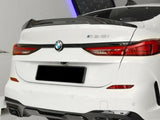 A white BMW 218i is parked indoors, showcasing its rear design with red taillights, dual exhausts, and carbon fiber accents. The setting is a sleek, modern space with bright lighting.