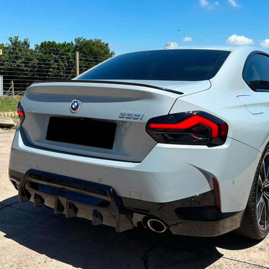 A silver BMW 220i car is parked on a concrete surface. It features red tail lights, dual exhausts, and text "220i" on the rear. Trees and a fence are visible in the background.