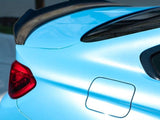 A light blue car's rear features a distinct black spoiler, red tail light, and fuel door. It is parked outdoors, with a blurred background of pavement and greenery.