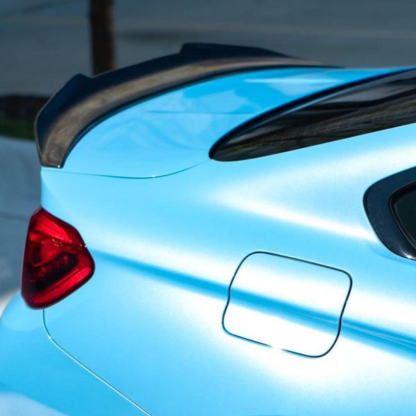 A light blue car's rear features a distinct black spoiler, red tail light, and fuel door. It is parked outdoors, with a blurred background of pavement and greenery.