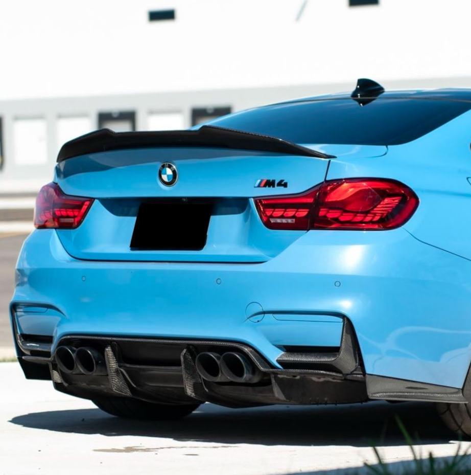 A blue BMW M4 car is parked in a sunlit area, showing its rear view with four exhaust pipes and a black spoiler. The BMW logo is visible, and the setting is urban.