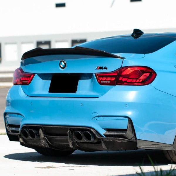 A blue BMW M4 car is parked in a sunlit area, showing its rear view with four exhaust pipes and a black spoiler. The BMW logo is visible, and the setting is urban.