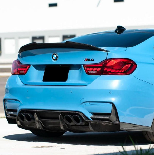 A blue BMW M4 car is parked in a sunlit area, showing its rear view with four exhaust pipes and a black spoiler. The BMW logo is visible, and the setting is urban.