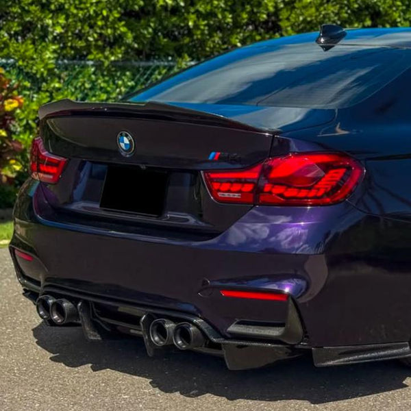 A dark purple BMW car with distinctive red taillights and dual exhausts is parked on a road, surrounded by green bushes and a clear sky reflecting on its surface.