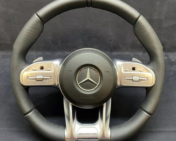 A black and silver steering wheel displays a prominent Mercedes logo at the center, surrounded by multifunctional buttons on the sides. "AIRBAG" and "AMG" are inscribed below.