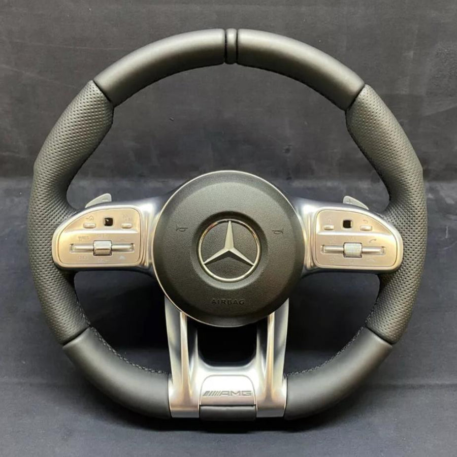 A black and silver steering wheel displays a prominent Mercedes logo at the center, surrounded by multifunctional buttons on the sides. "AIRBAG" and "AMG" are inscribed below.