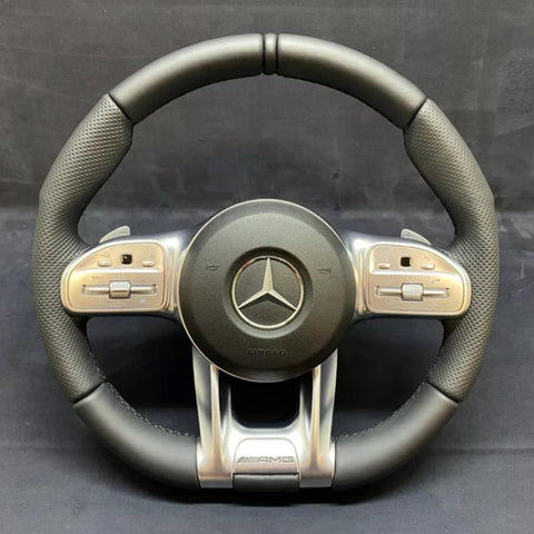 A black and silver steering wheel displays a prominent Mercedes logo at the center, surrounded by multifunctional buttons on the sides. "AIRBAG" and "AMG" are inscribed below.