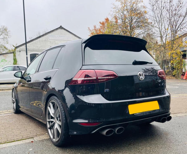 A black hatchback car is parked on a street near autumn trees and a warehouse. It features dual exhausts and alloy wheels, with a visible "VW" logo on the rear.