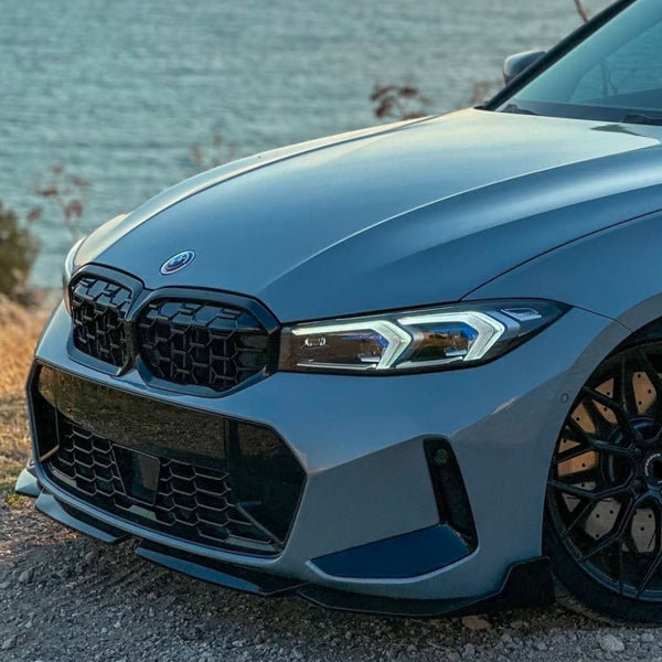 A sleek, silver car with distinct headlights and a prominent grille is parked on a rocky surface, overlooking a body of water with a few scattered plants nearby.
