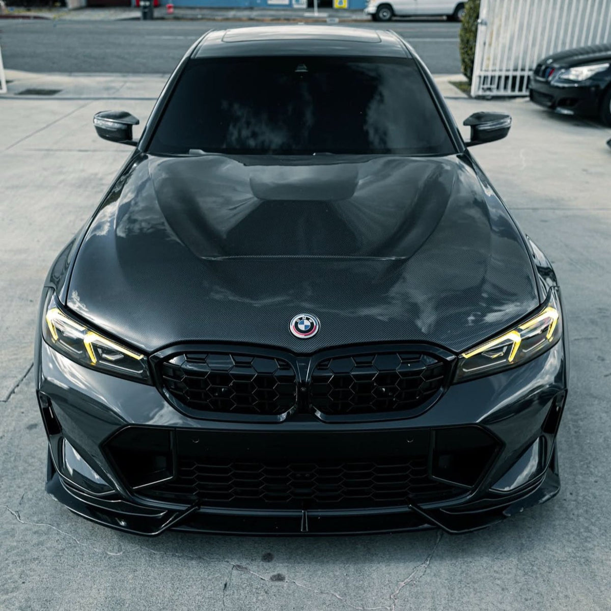 A sleek black BMW with tinted windows is parked on a concrete driveway. It features prominent yellow headlights and a shiny grille. Background includes a fence and parked cars.