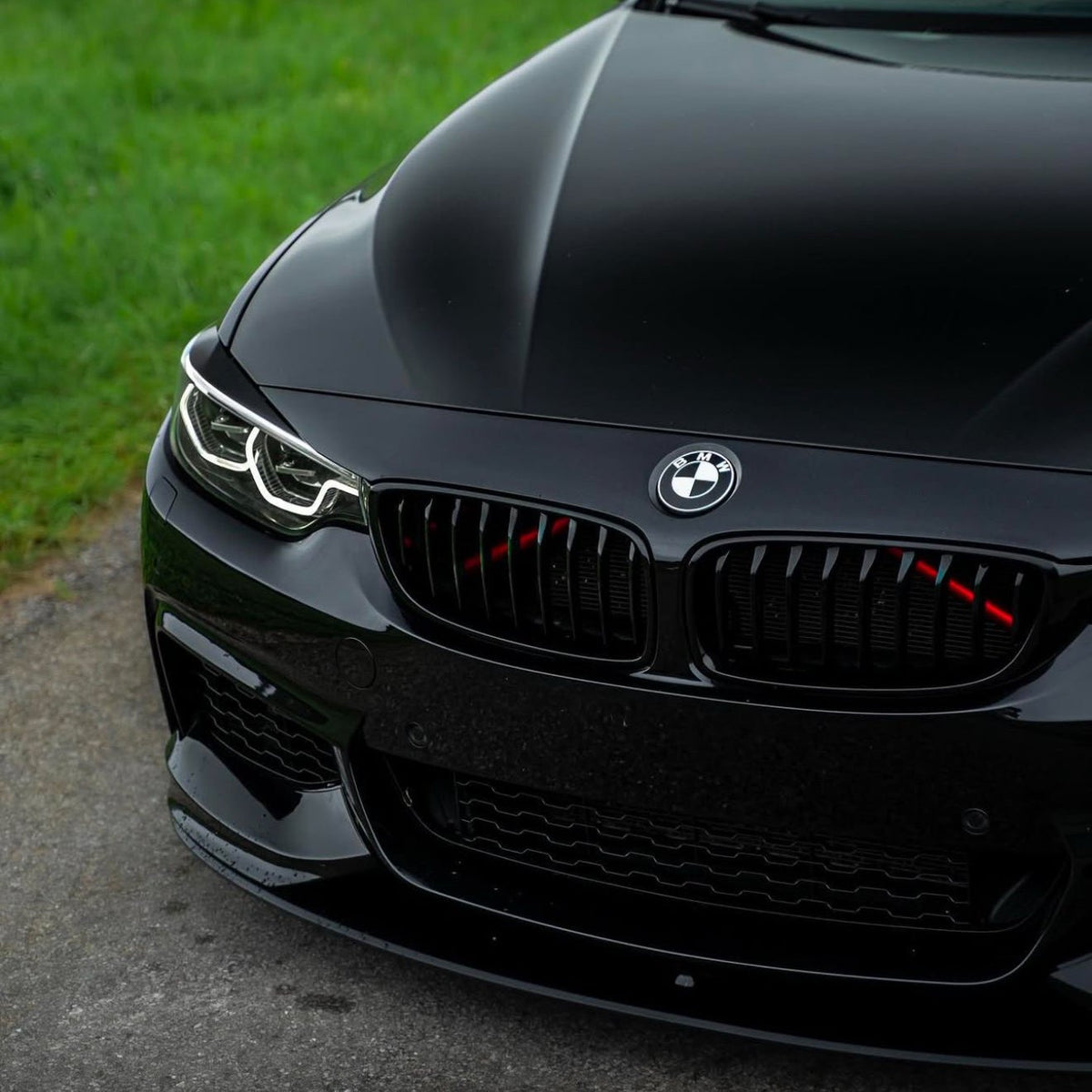 A black car with a BMW emblem on the hood is parked on a pavement beside green grass. Its headlights and grille are prominently featured, displaying a sleek, modern design.