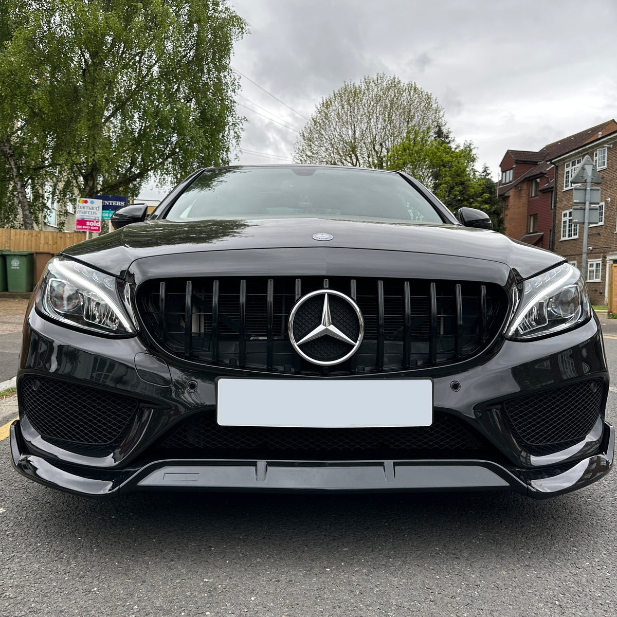 A black Mercedes-Benz car is parked on a suburban street near brick houses and green trees, with a real estate sign in the background.