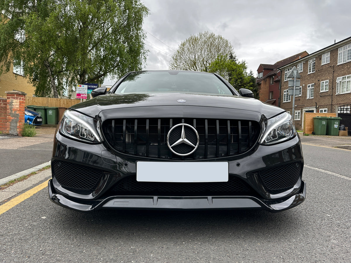 A black Mercedes-Benz car is parked on a suburban street near brick houses and green trees, with a real estate sign in the background.