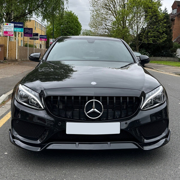A sleek black car, parked on a suburban street, features a prominent Mercedes-Benz logo on the grille. Surrounding are trees and buildings with real estate signs visible in the background.