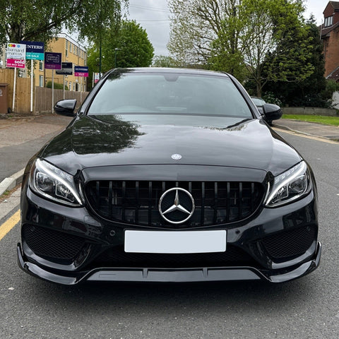 A sleek black car, parked on a suburban street, features a prominent Mercedes-Benz logo on the grille. Surrounding are trees and buildings with real estate signs visible in the background.