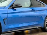 A blue car is parked on wet pavement, showcasing its sleek side profile and glossy finish. It features the M Performance logo near the bottom. The background includes a brick wall.