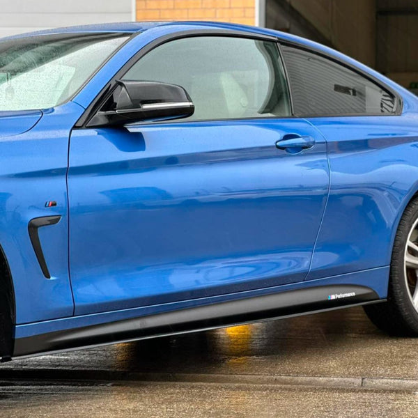 A blue car is parked on wet pavement, showcasing its sleek side profile and glossy finish. It features the M Performance logo near the bottom. The background includes a brick wall.
