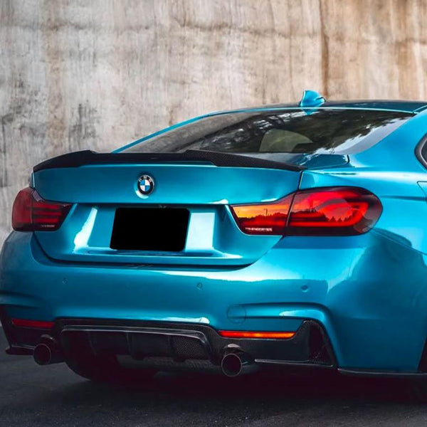 A blue BMW car is parked beside a concrete wall, showing its rear end with dual exhaust pipes and the BMW logo centered on the trunk.