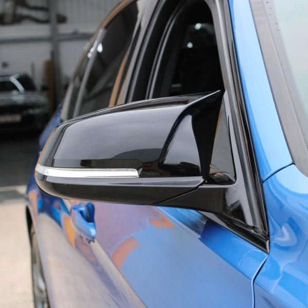 A sleek black side mirror is attached to a blue vehicle, reflecting its surroundings. The car is parked in an indoor area with concrete flooring and other vehicles visible in the background.