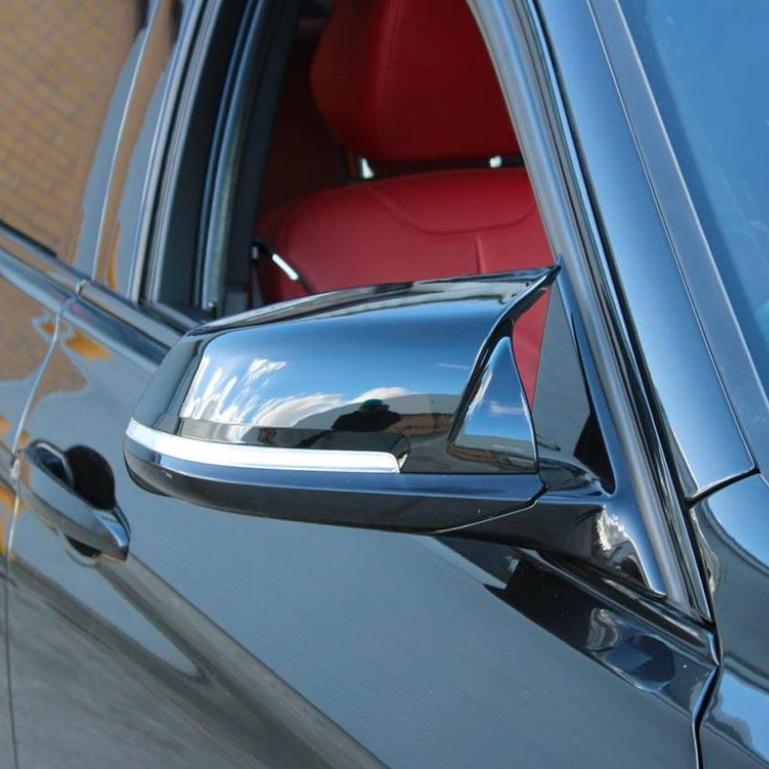 A sleek black car's side mirror reflects the sky and surroundings. The open window reveals red interior seats, set against an urban environment.