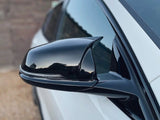 A sleek black side mirror on a white car reflects the surroundings, set against a blurred, rustic outdoor backdrop.