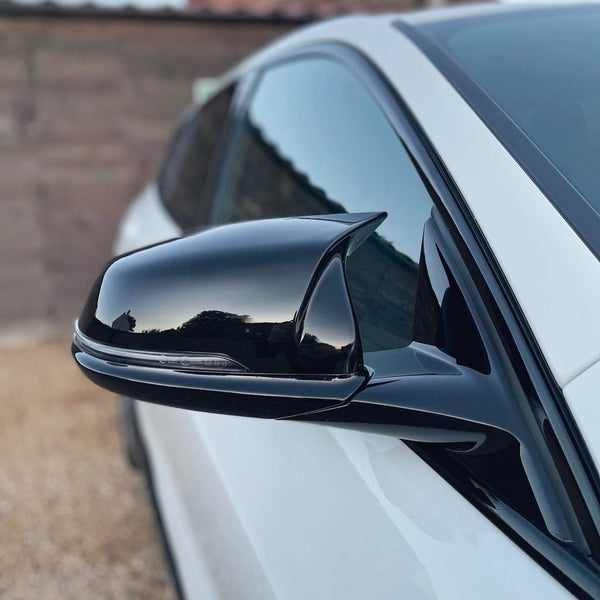 A black side mirror reflects the surroundings on a sleek white car, parked on a gravel driveway with a wooden fence in the background.