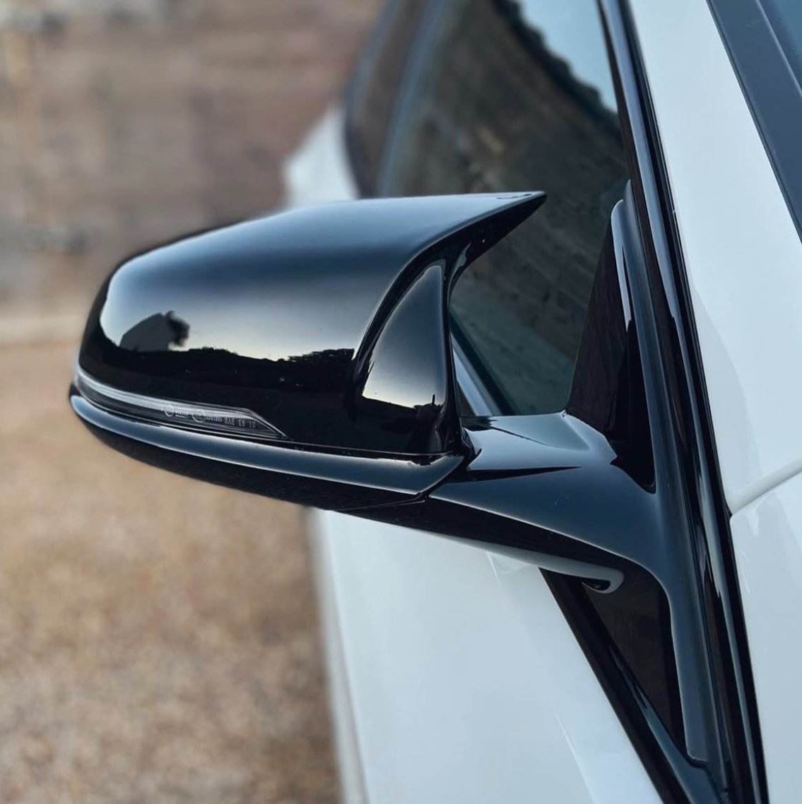 A sleek black side mirror is attached to a white car, reflecting a blurred outdoor scene, with a gravel ground visible in the background.