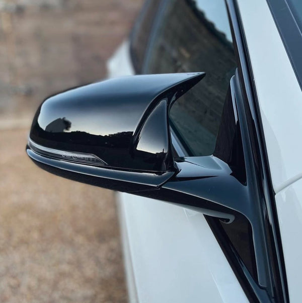 A sleek black side mirror is attached to a white car, reflecting a blurred outdoor scene, with a gravel ground visible in the background.