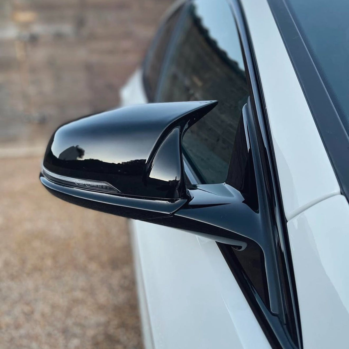 A sleek black car side mirror reflects surroundings, attached to a white vehicle parked outdoors on a gravel surface with a blurred wooden backdrop.