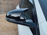 A sleek black car side mirror reflects the sky, attached to a white vehicle. The background is a blurred, neutral-toned wall.