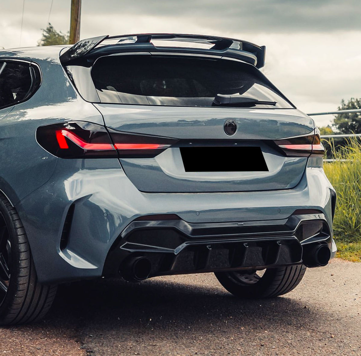 Sleek car parked on a road, displaying sporty rear design and glossy finish. Red taillights highlight its modern contours against a background of greenery and cloudy sky.