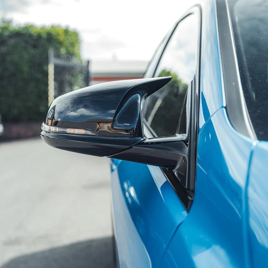 A black side mirror is attached to a blue car, reflecting the sky and surrounding greenery; the scene is outdoors in a paved area with a fence in the background.