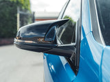 Side mirror reflecting sky, attached to a blue car. The background shows a blurred fence and greenery under partly cloudy skies.