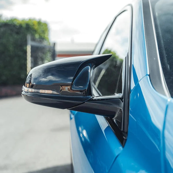 A shiny black side mirror is attached to a bright blue car, reflecting a cloudy sky. The vehicle is parked near a hedge and gate under a partly cloudy sky.
